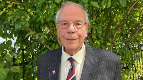 Francis Wakem wearing a white shirt, red blue and white striped tie and grey pinstripe blazer. He is wearing a Victim Support pin on his lapel and is smiling at the camera, standing in front of a tree.