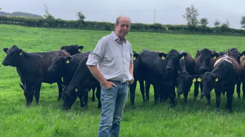 Farmer Ian Buchannan pictured successful  a airy  white-coloured  garment  and jeans portion    lasting  successful  beforehand   of a fig   of cows successful  a field. 