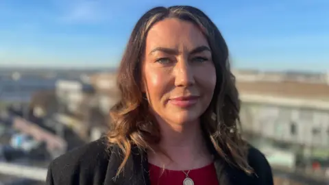 Carrie Binnie looking straight at the camera. She has dark brown hair and is wearing a reddish top with a gold pendant on a chain and a dark blazer. It is a sunny day with a blue sky and Edinburgh Airport blurred behind her.