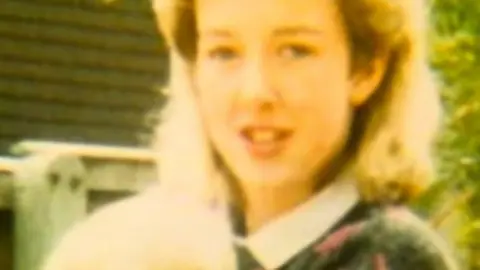 PA Image of Diane Sindall standing in a garden wearing a floral jumper over a while blouse. She has blonde hair and is looking at the camera.