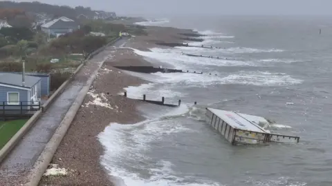 Eddie Mitchell A shipping container floats in a stormy, grey sea. It is right next to the coast. 