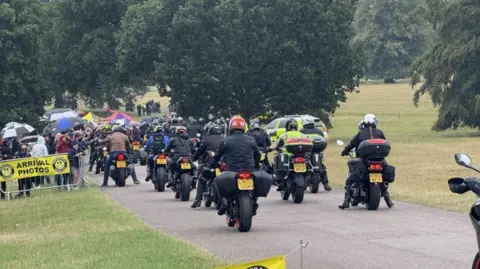 A convoy of motorcyclists seen from behind coming to a stop on a narrow path in a country park, surrounded by grass and trees.