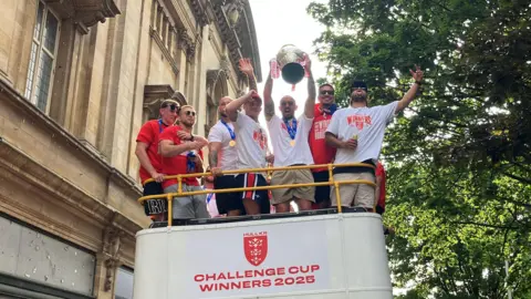 Seven players, dressed in red and white t-shirts, stand on top of a white open top bus with the rugby league Challenge Cup.
