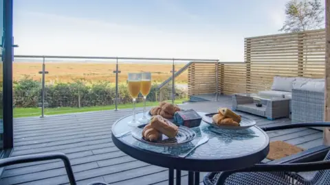 John Phillips The images shows a glass table on the balcony overlooking the Gower, there are champagne flutes and pastries on the table and a view to a large flat stretch of countryside beyond. 