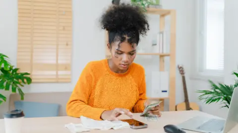 Getty Images A young woman with dark hair tied up wearing an orange jumper holding cash with her phone, bills and laptop around her.