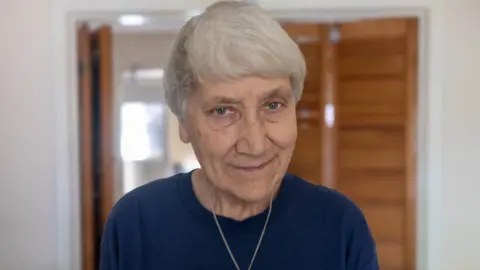 Mary Joy Langdon, 76, smiling for the camera in an interior room. She is wearing a dark-coloured jumper.
