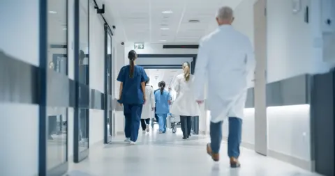 Getty Images Female and male healthcare officials walk down corridor with their backs to the camera.