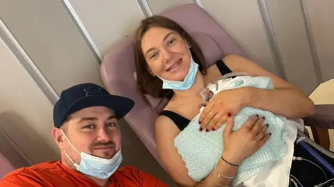 Laura Pridding smiles at the camera, she has long brown hair and has a face mask around her chin. She is holding her newborn baby George in her arms, he is wrapped in a blue knitted blanket and there are tubes going beneath the blanket. Next to them is dad Ali Davies who is wearing a black cap and an orange t-shirt. He also has a facemask below his chin. 