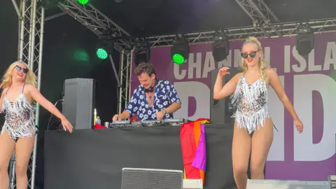 A stage performance at the Channel Islands Pride event features two dancers in sparkly fringed outfits on either side of the stage and a DJ in the centre wearing a blue floral shirt. The DJ stands behind a table with equipment, draped with a rainbow flag. A purple banner in the background displays partially visible white text reading 'CHANNEL ISLANDS PRIDE,' with green stage lights shining above.