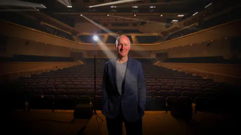 A man standing on a theatre stage beneath bright spotlights, hands in his pockets, with rows of empty seats and a large auditorium stretching out behind him.