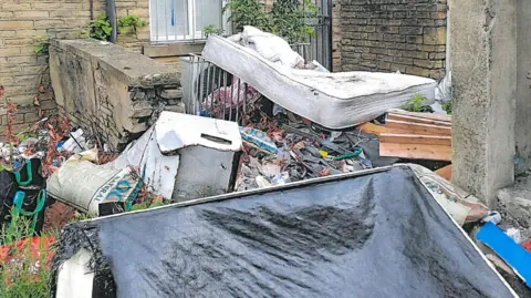 A neglected outdoor yard filled with dumped household waste and furniture. Several mattresses, broken boards, bags, and miscellaneous rubbish are piled together among weeds and fallen leaves. The area is enclosed by worn stone and brick walls, with a barred window visible in the background.
