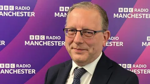Michael Winstanley smiles while standing next to some BBC Radio Manchester branding. 