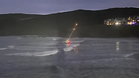 Chris Parsons A small yacht with lights on the mast and hull lies at an angle on a beach as it gets dark with sea and beach visible below a row of houses with lights on.