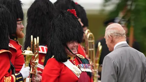 PA Media King Charles III (right) during the Ceremony of the Keys in the gardens of the Palace of Holyroodhouse in Edinburgh. He has his back to the camera. Facing the camera are members of the band of the Royal Regiment of Scotland. They are wearing red and gold jackets, a black feather bonnet and are carrying gold instruments.