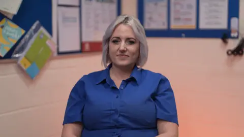 Carly-Ann has platinum blond hair in a chin-length bob with a centre parting. She is wearing a blue, short-sleeved shirt - the uniform of the Lincolnshire Co-op. She is seated in a staff room, with blue noticeboards behind her.