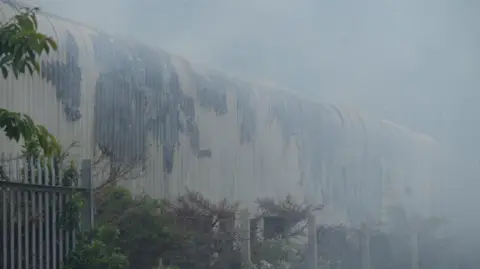 Richard Knights/BBC Burnt parts of the roof and building has been left behind amongst a plume of smoke at the warehouse.
