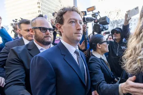Meta boss Mark Zuckerberg arrives at an LA court wearing a dark blue suit jacket, white shirt and grey tie. He is surrounded by security men