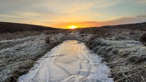 A frozen lochan surrounded by frozen glass with a low winter sun peeping over the horizon