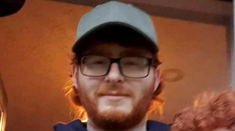 Photo of Joe Scott. He has a ginger beard and hair, and is wearing glasses and a cap
