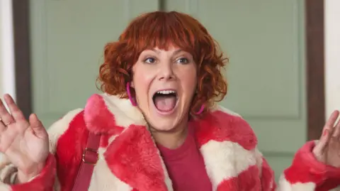 BBC/Expectation TV/Neil Sherwood Sophie Willan starring in series two of Alma's Not Normal - she has red short curly hair and a pink and white striped fake fur coat