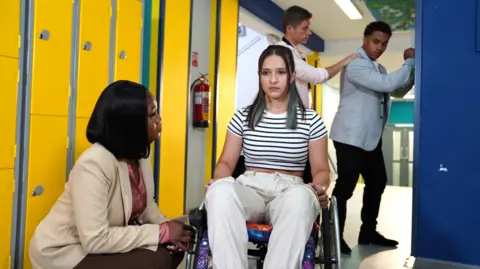 Lime Pictures Ellie sits in a wheelchair, looking serious. She is in a school corridor next to a set of yellow lockers. A woman is crouching next to her and looking at her, while in the background a teenage boy leans against more lockers and a teacher puts a hand on his shoulder.