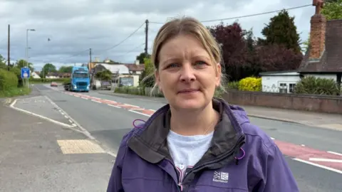Rebecca Hill is wearing a purple Karrimor water proof and a grey T-shirt. She has blonde hair, which is tied back. She also has a heart-shaped pendant. Behind her is a main road, on which a light-blue lorry and a red car are travelling.