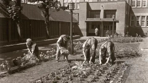 Borthwick Institute/Heritage Images/Getty Images Gardeners planting a bedding display. A black and white photo with four gardeners in the foreground and the factory building in the background.