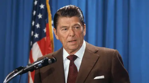 Getty Images Uma foto de arquivo de Ronald Reagan da década de 1970. Ele usa um paletó marrom, gravata vermelha escura, camisa branca e cabelo castanho penteado para trás. Ele fala ao microfone e fica diante de uma bandeira americana e uma tela azul.