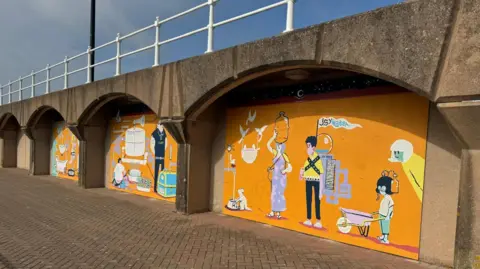 BBC Three murals on the Elizabeth Marina in Jersey, orange backgrounds with cartoon people, railings above, grey and blue skies