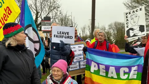 A group of protesters holding signs. One has a rainbow flag with the word "peace", others have simpler signs such as one saying "leave Fairford". It is a grey day.