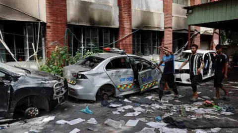 Reuters People walk next to vandalised cars at the Mohammadpur Police Station, after the resignation of Bangladeshi Prime Minister Sheikh Hasina, in Dhaka, Bangladesh, August 6, 2024.