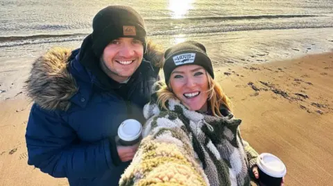 Andy Johnson Photograph of Andy and Danni Johnson, stood on a beach with the sun reflecting on the water behind them. They both hold takeaway coffee cups and smile towards the camera. Andy (left) wears a black beanie and a navy puffer coat, while Danni Johnson wears a fleece jacket and black beanie. 