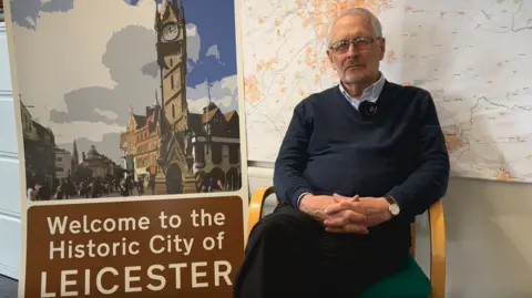 A man sits in a chair next to a brown sign that reads Welcome to the Historic City of Leicester
