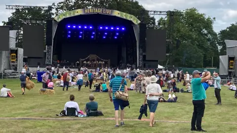 Jill Bennett/BBC People in front of an outdoor stage sitting on grass or walking. There is a "Heritage Live" logo above the stage.  There are 20 blue lights in a pattern on the backdrop of the stage. There are large black speakers on either side of the stage.