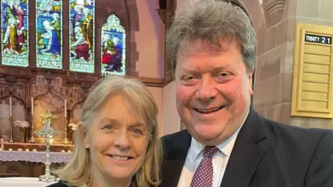 Sue and Alex Tatham inside the church with a stained-glass window and candles behind them.