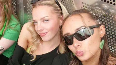 Two girls in black tops. The one on the left has blonde hair, and the one on the right has brown hair. They wear sunglasses.