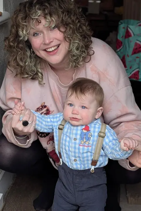 Victoria Cain A mum with curly blonde hair smiles as she guides her infant son to help him take steps. The baby boy wears a Paddington bear blue checked shirt with braces and blue trousers, and the mum wears a pink sweatshirt.