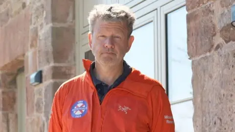 Matt Neilson looks at the camera. He has short thick fair hair, very faint stubble and is wearing an orange mountain rescue jacket. He is standing in front of a stone house he is building.