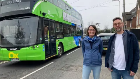Two people are standing the right of the picture. A man and a woman. To their left is a green bus.