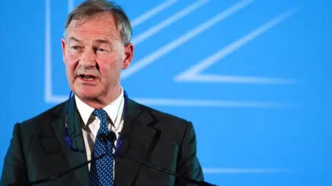 Rupert Lowe is wearing a dark business suit, a white shirt, patterned blue tie and has a pair of glasses hanging round his neck. He is standing at a podium with a microphone. The background is a bright blue presentation screen with abstract white geometric lines.