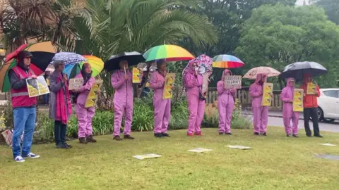 A line of 10 protestors, many in pink hazmat suits holding umbrellas and placards