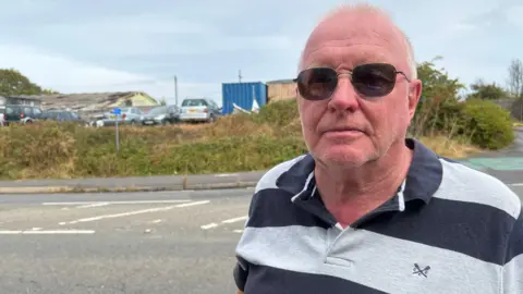 Martin Fey pictured at Locking Farm Industrial Estate in Weston-super-Mare. He has short white hair and is wearing dark sunglasses and a grey and navy blue striped shirt. 