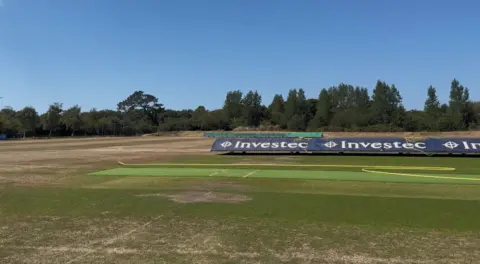 A green grass pitch with a yellow hose and a cover to go over the cricket wicket.