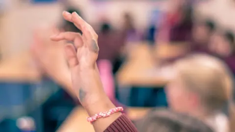Eric Johnson A child's hand is raised in a classroom. There's some black ink on the hand and the background is blurred.