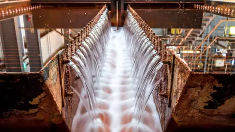Water pours from a series of outlets in two rows into a stream of water in the middle. On the other side of the two rusty walls are two flat bodies of water. 