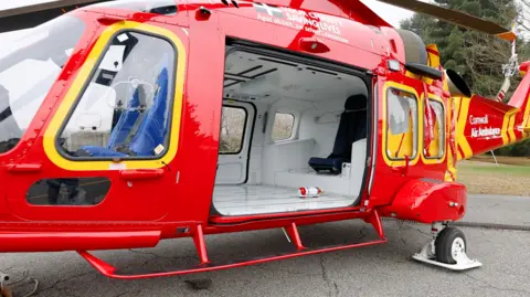 A red helicopter with the logo Cornwall Air Ambulance on its tail has landed on the ground.
An open door shows that the inside is an empty shell before fitting out with medical equipment.