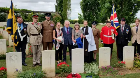 MOD Crown Eleven people dressed in various military uniforms with two holding flags stand next to a series of grey headstones with flowers and poppy wreaths at their base.