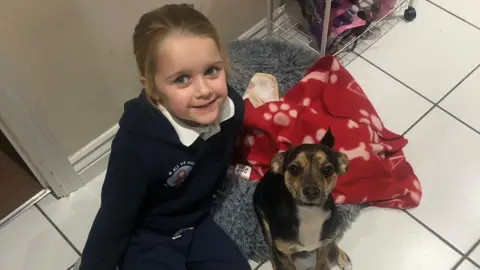 Lauren Marsh Bonnie Marsh is sitting on a white tiled floor with a brown and black small dog. There is a red blanket and a grey dog bed on the floor.