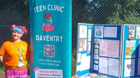 Daventry Town Council A woman with multicoloured hair tied in bunches is standing next to a banner, wearing an orange T-shirt and purple skirt. The roll-up banner is blue with the words "TEEN CLINIC DAVENTRY" and a QR Code. There is a noticeboard to the right of the banner, showing various pictures and notices. There is metal mesh in the background with a hedge behind