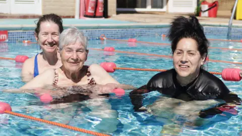 RUH Three women, including an elderly lady, in an outdoor swimming pool, all smiling to camera in the sunshine. There are lanes set up with orange ropes with small pink floats. 